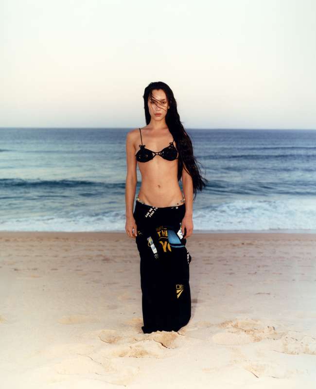 A woman in a bikini and a wrap skirt stands on a beach with the ocean and sky in the background at dusk, in a medium shot.