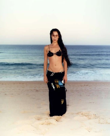 A woman in a bikini and a wrap skirt stands on a beach with the ocean and sky in the background at dusk, in a medium shot.
