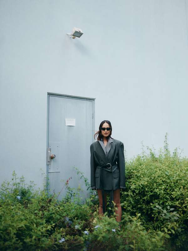 A stylish woman wearing a belted gray blazer stands in front of a muted blue door in a lush overgrown outdoor setting.