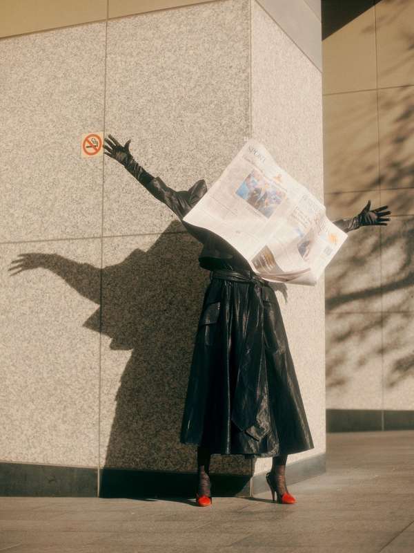 A fashion shot of a figure in a black skirt, gloves, and red heels, obscured by a newspaper, against a light-colored building.