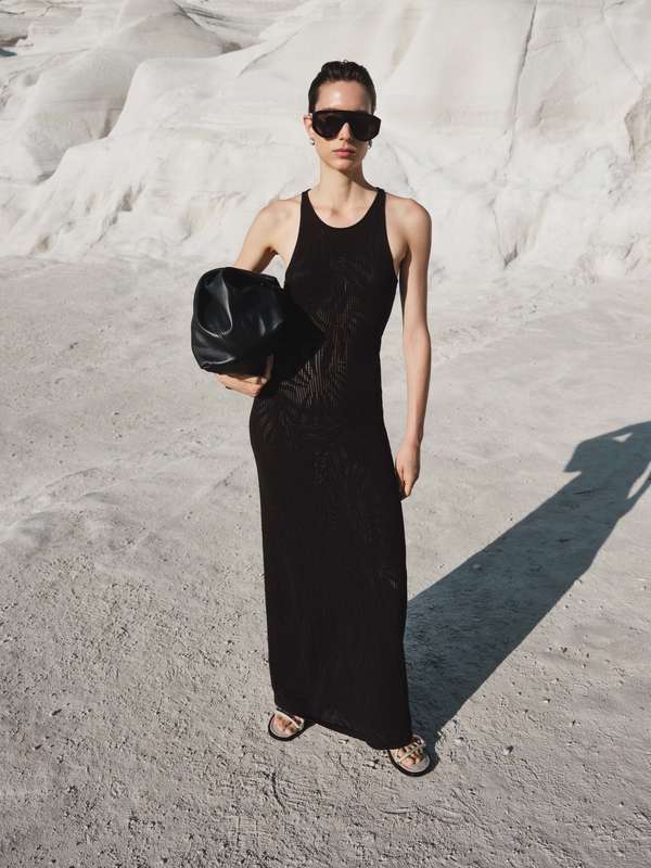 A slender adult woman in a black maxi dress and sunglasses holds a black bag, posing in an outdoor setting with white rock formations.
