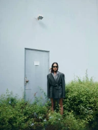 A woman wearing dark sunglasses and an oversized dark green belted blazer standing outside against a pale wall.