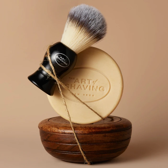 Shaving brush and round soap cake stacked on a dark wooden bowl against a warm beige background.