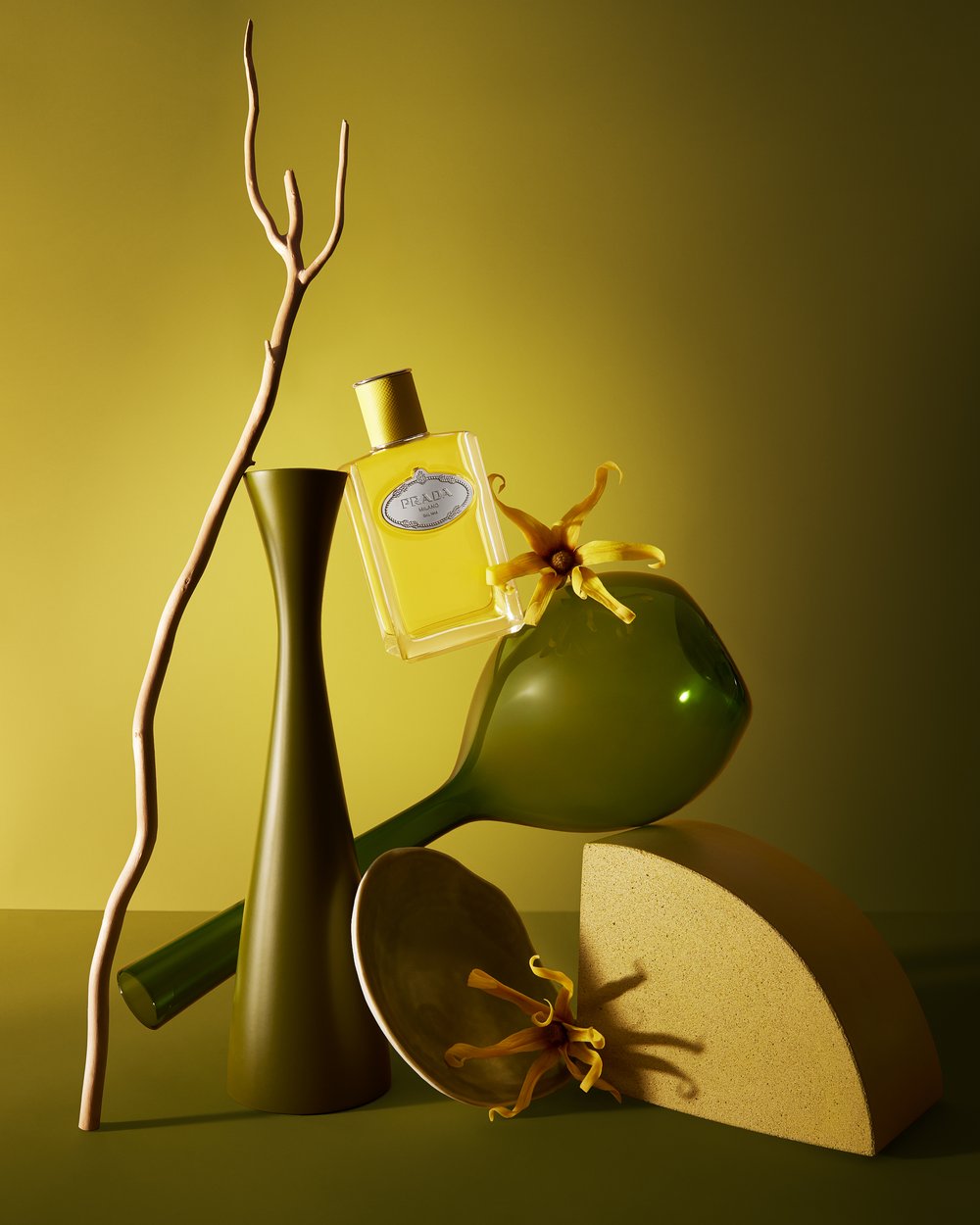 Still life featuring a Prada perfume bottle, a green vase, flowers, a balloon, and a geometric shape against a yellow-green background.