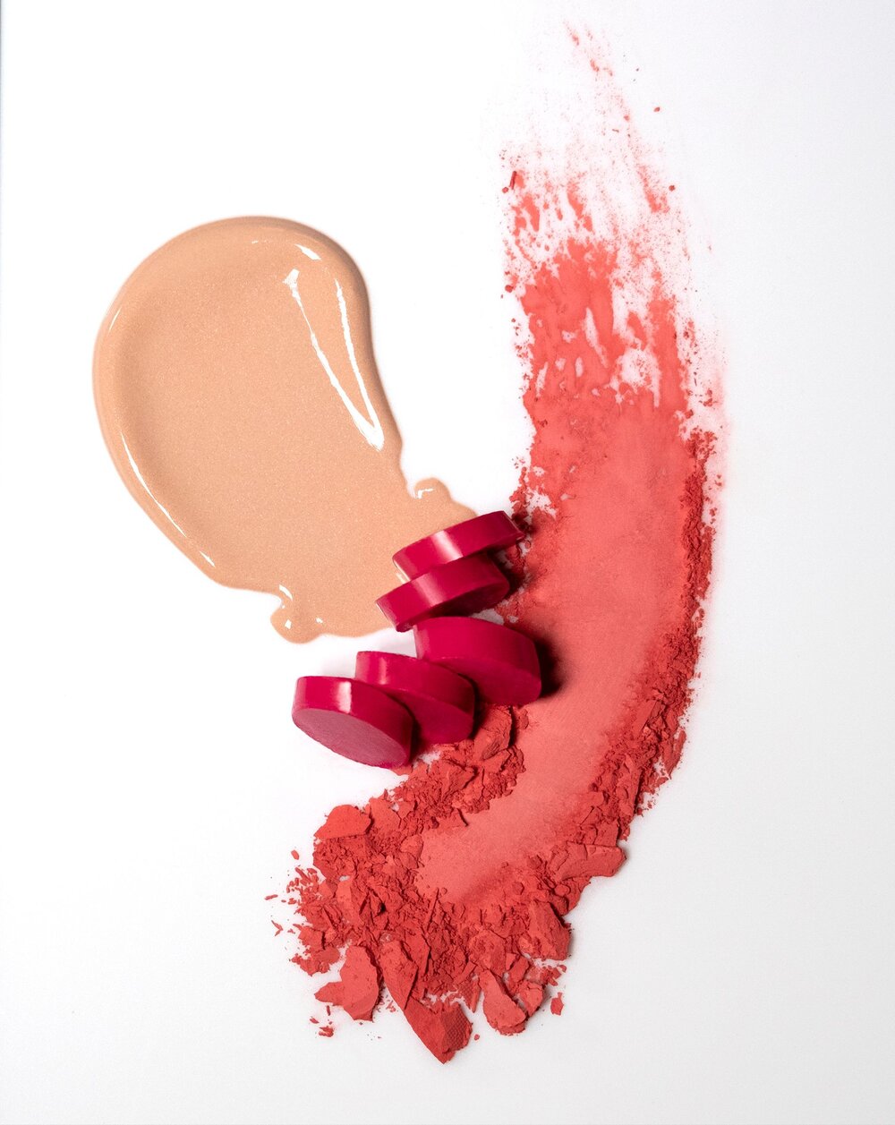 Close-up of red lipstick, beige foundation, and coral powder makeup swatches on a white surface, showcased in a beauty still life.