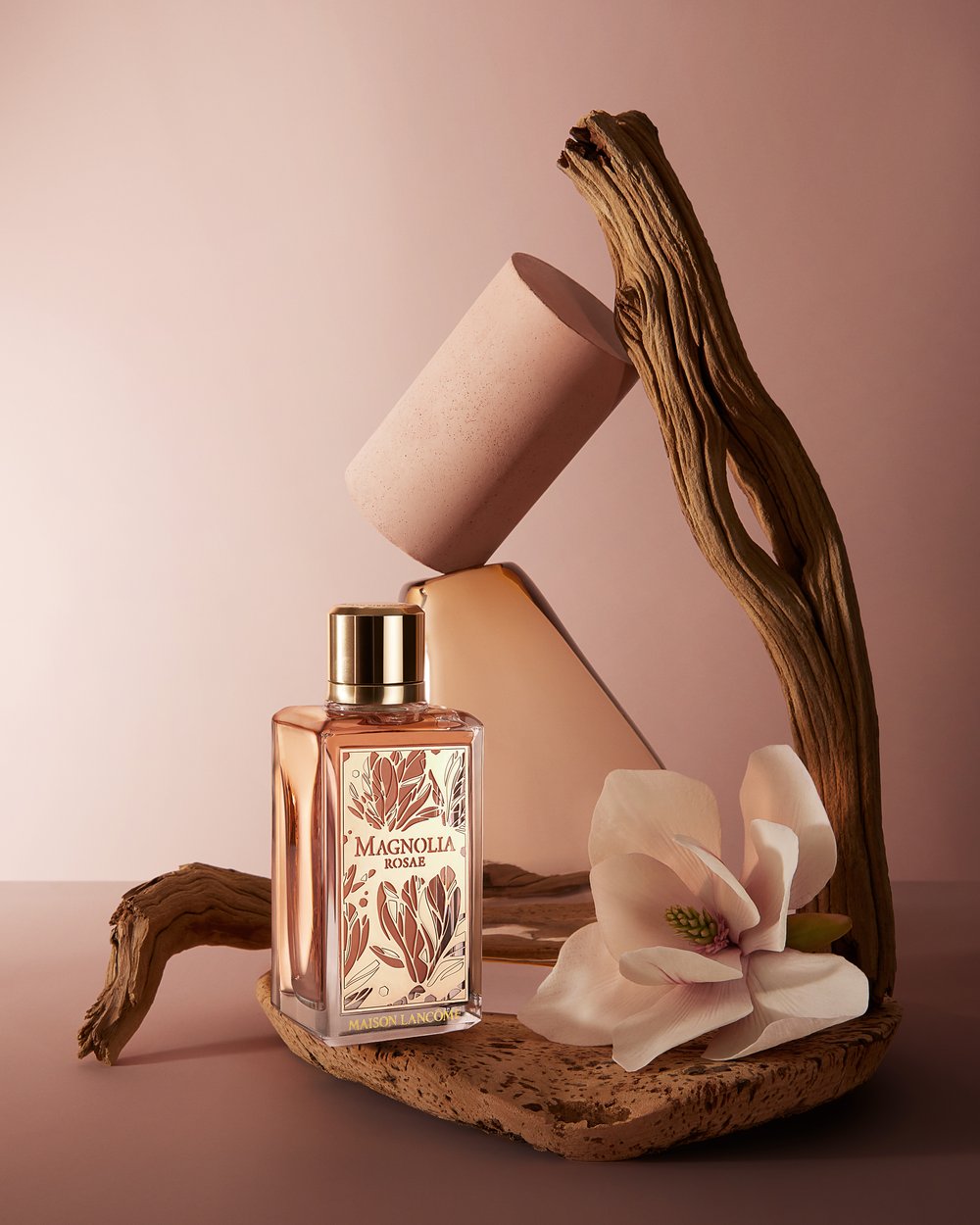 Perfume bottle of Magnolia Rosae, a cylinder, a flower, and a piece of driftwood arranged on a pink surface against a pink background.