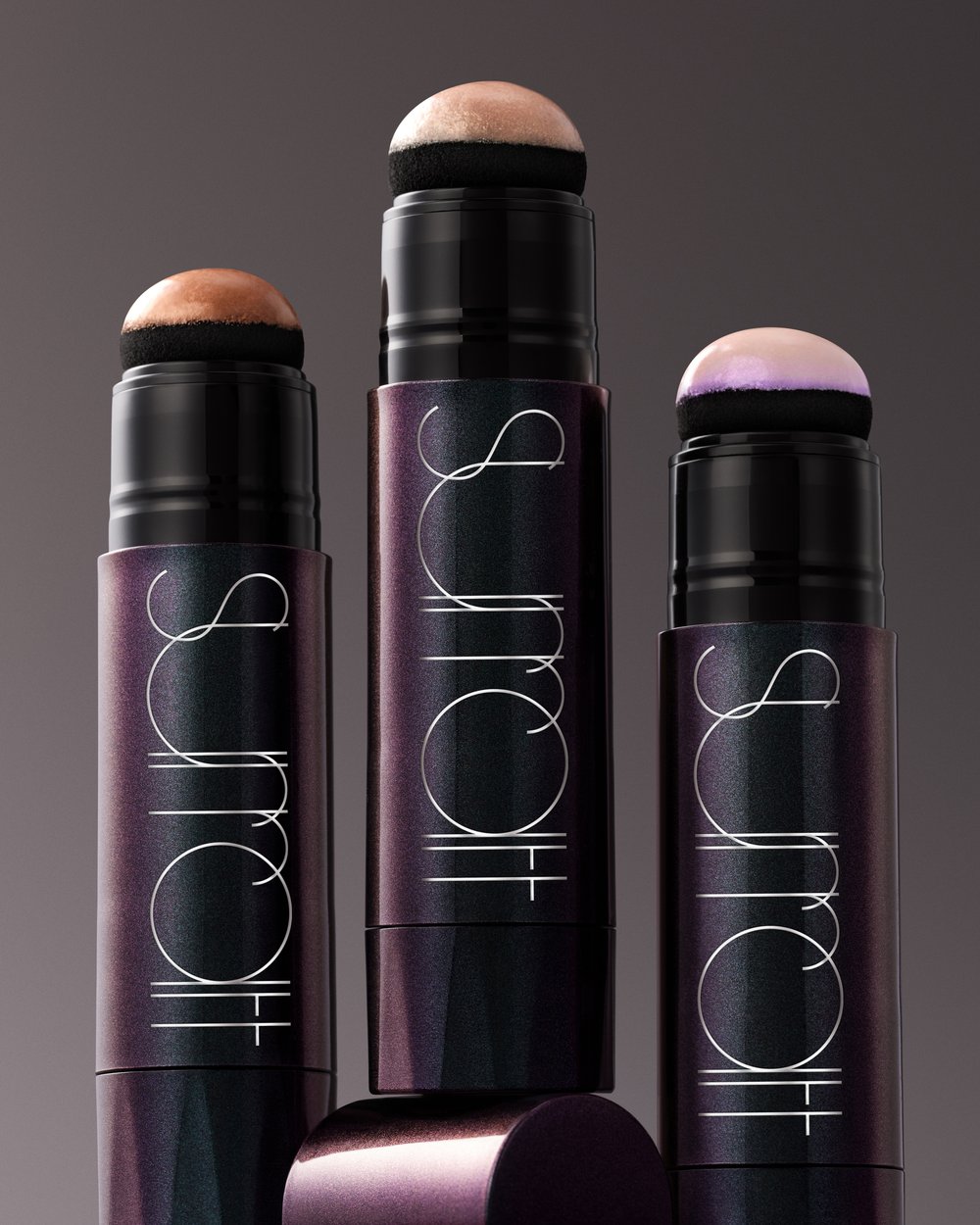 Three makeup sticks with applicator sponges, showcasing different color variations, against a gradient grey background in a studio shot.