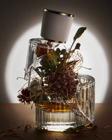 Still life of a perfume bottle balanced on broken glass and dried flowers, with soft lighting creating a spotlight effect.