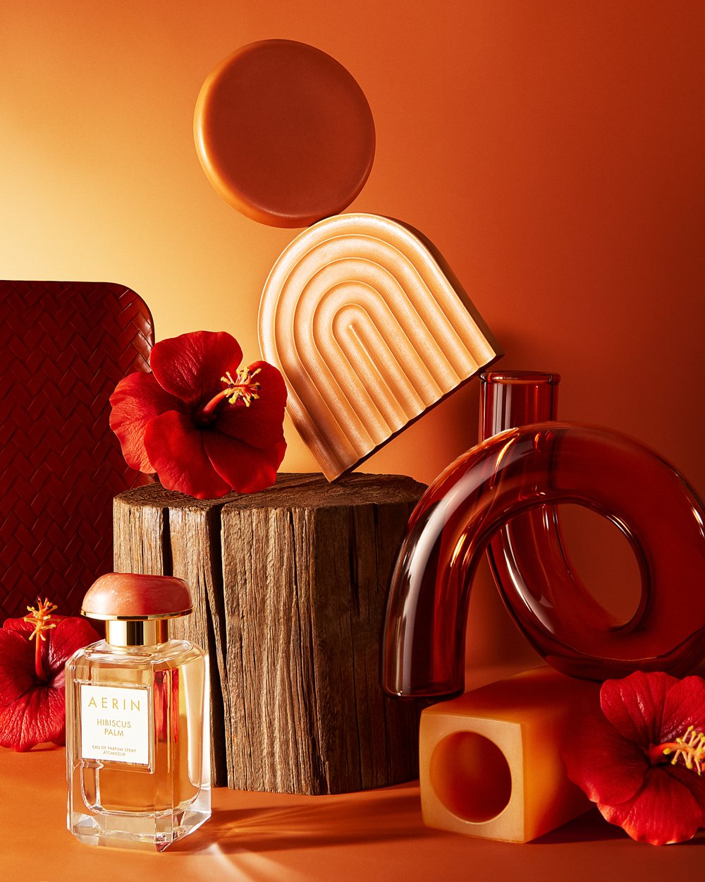 Still life shot of a perfume bottle, red hibiscus flowers, and geometric objects in brown and orange hues against a gradient background.