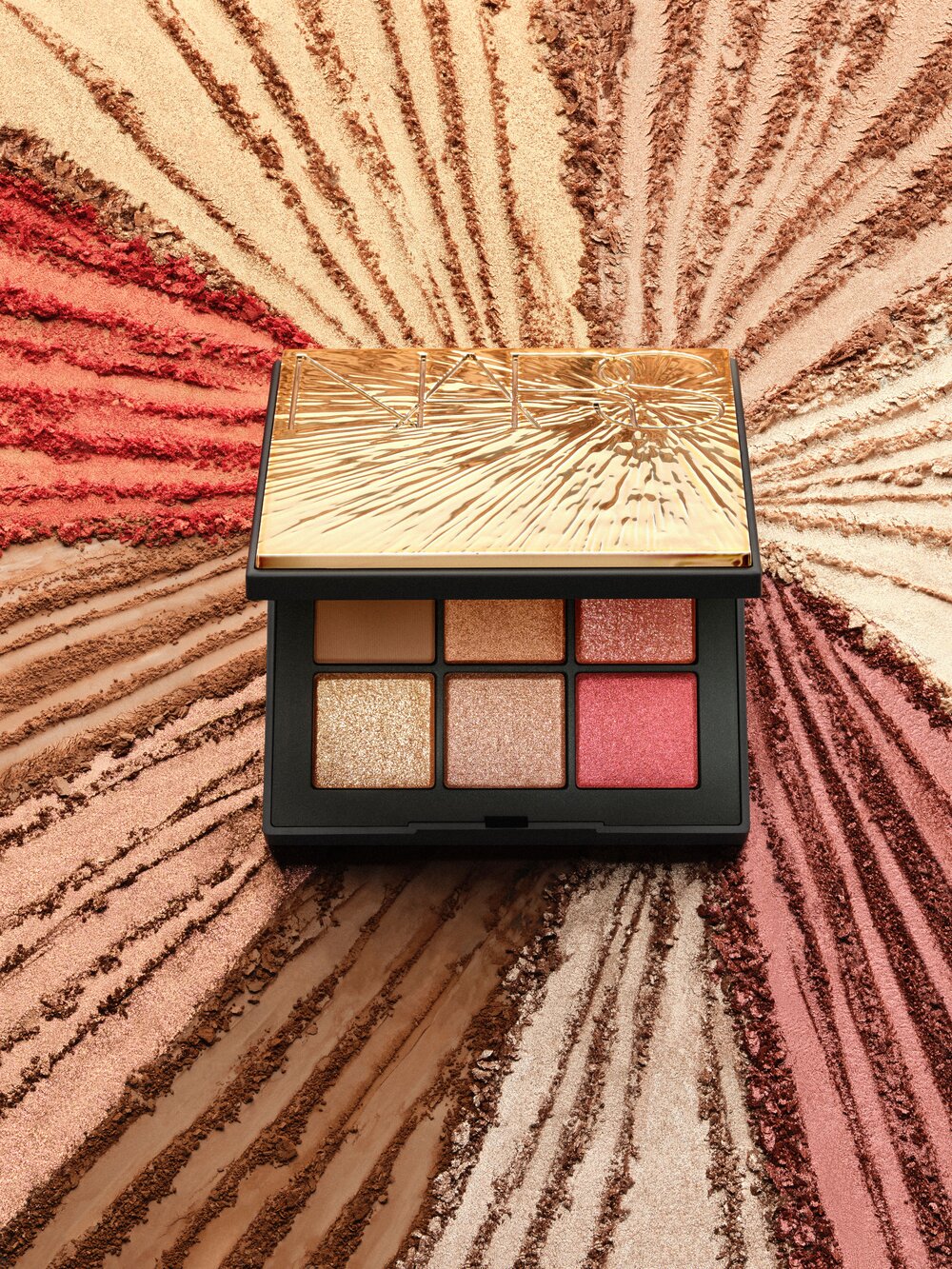 Makeup palette featuring gold, peach, and pink eyeshadows, set against a background dusted with loose powder in radiating patterns.