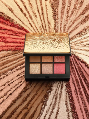 Makeup palette featuring gold, peach, and pink eyeshadows, set against a background dusted with loose powder in radiating patterns.