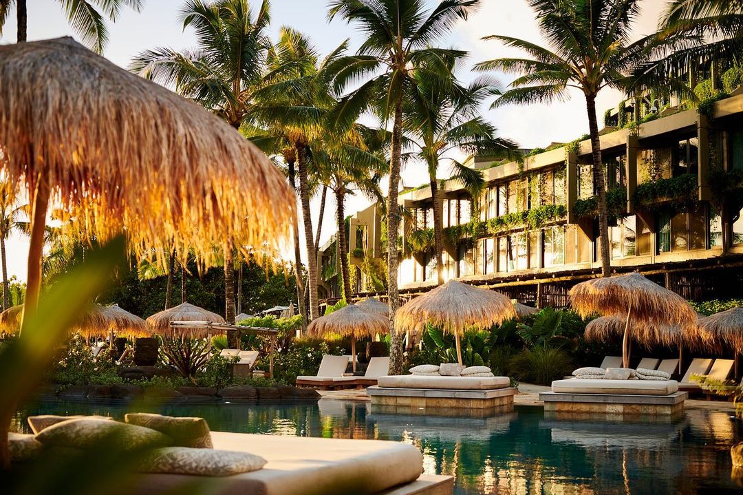 Lounge chairs and umbrellas around a tropical pool at luxury resort in Hawaii with palm trees and hotel building during daytime.