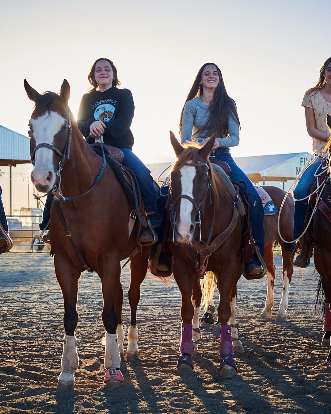 Three young women riding horses outdoors in arena. Horses are brown with white markings, wearing riding gear in a full shot.