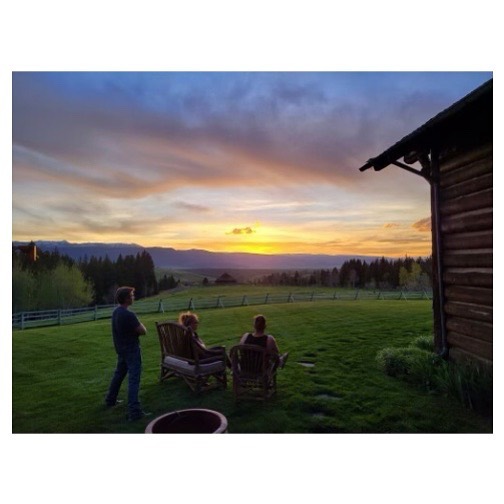 People relaxing outdoors on a lush green lawn at sunset, enjoying the scenic mountain view from a wooden cabin's vantage point.