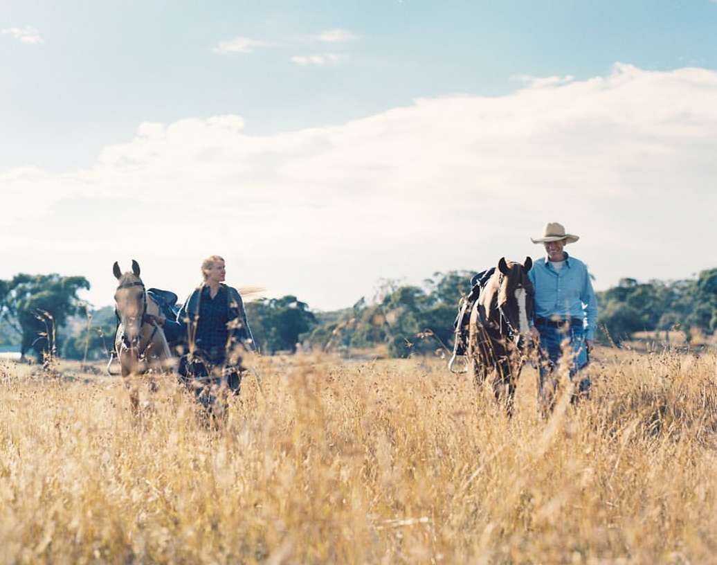 Two people walking with horses through a field of tall, dry grass under a partly cloudy sky, creating a peaceful, rural scene.