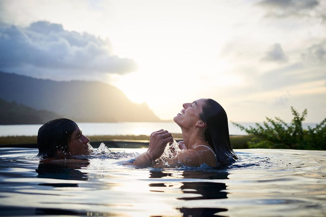 A couple laughs and splashes in a reflective infinity pool with a distant mountain backdrop against a sunset sky with soft, warm light.