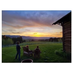 Three people watching sunset over grassy meadow and distant mountains from wooden cabin porch
