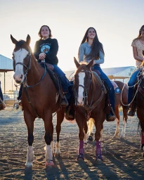 Three smiling young women horseback riding in an outdoor dirt arena during golden hour, photographed by Mikkel Vang.