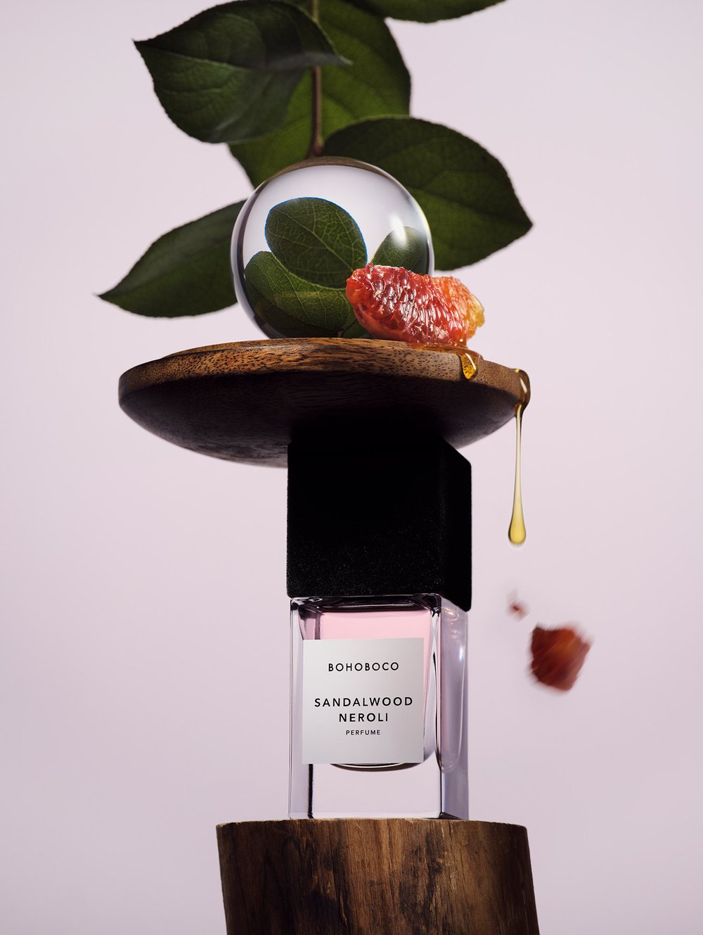 Still life composition featuring a Bohoboco Sandalwood Neroli perfume bottle balanced on wood, complemented by greenery and citrus.