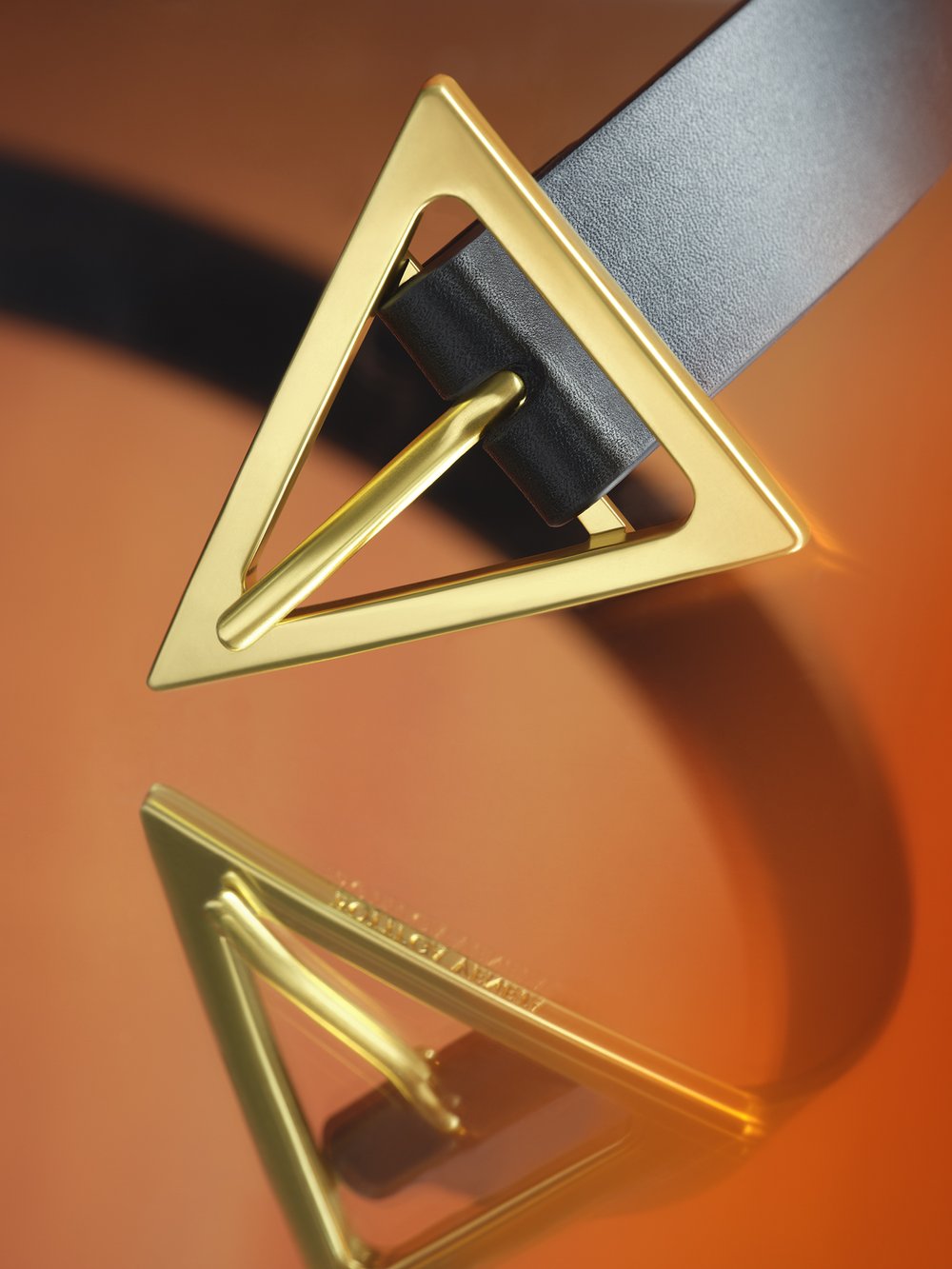 Close-up of a black leather belt with a gold triangular buckle on an orange reflective surface, showcasing luxury and design.