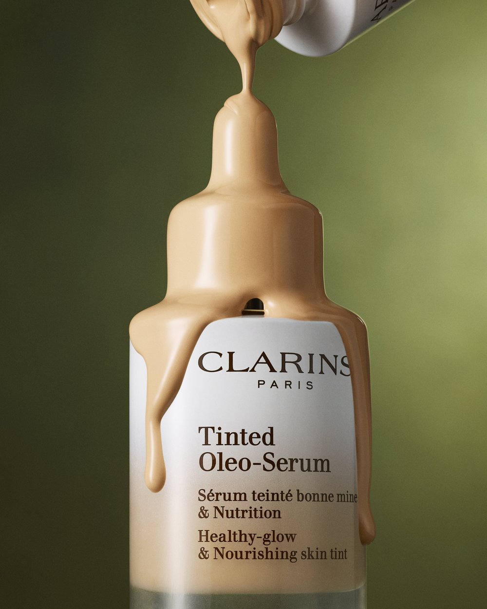 Clarins Tinted Oleo-Serum bottle with beige liquid foundation product being poured on it, against a soft green background.