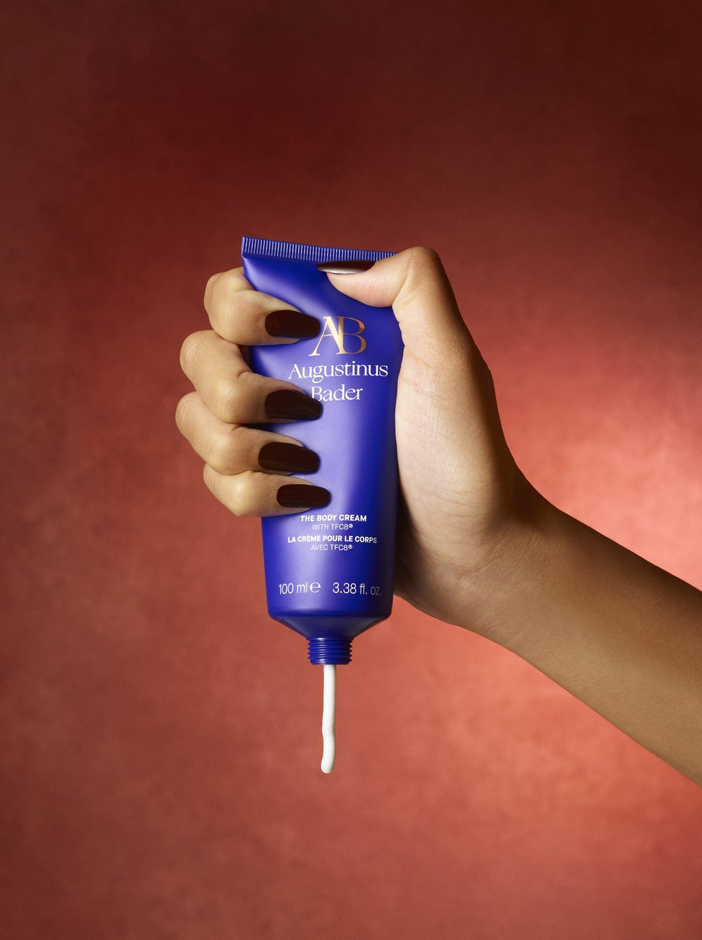 Hand holding blue Augustinus Bader body cream tube against a textured, dark red background with product dripping from the tube.