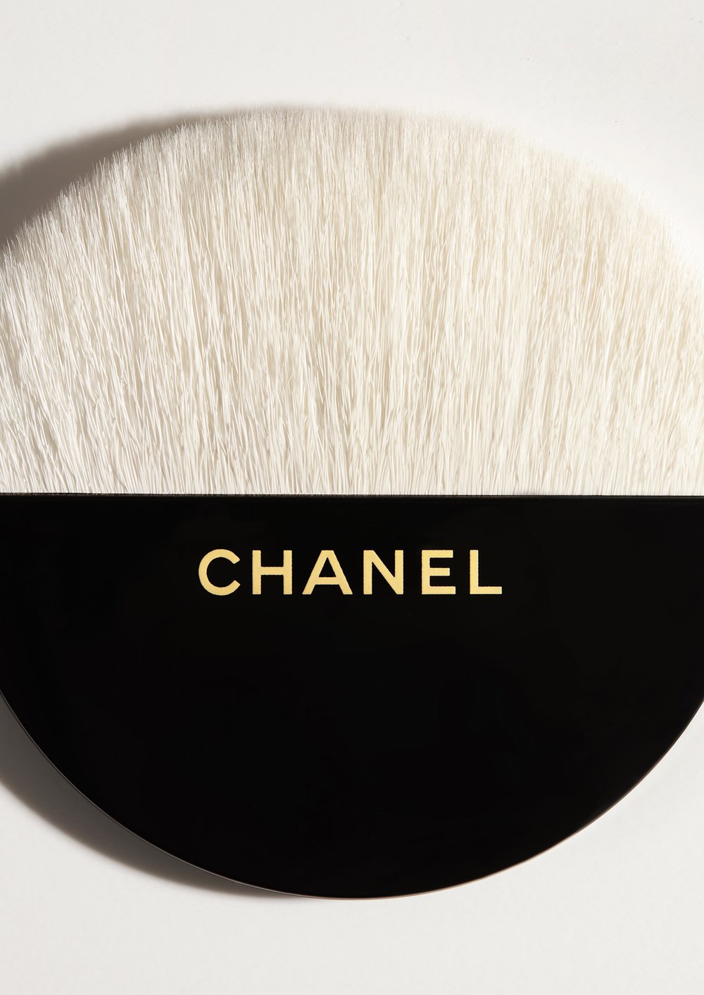 Close-up studio shot of a makeup brush from beauty brand Chanel with a black handle and soft white bristles against a white background.