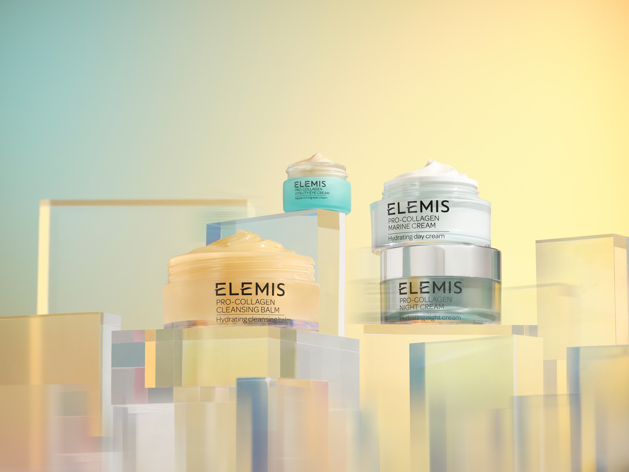 Cosmetic product jars from Elemis displayed on transparent geometric shapes with a light blue and yellow gradient background. Beauty concept.