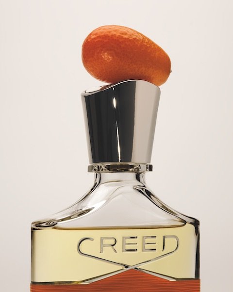 Perfume bottle with silver cap and orange citrus fruit on top against a white background in a studio product shot.