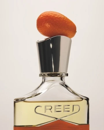 Perfume bottle with silver cap and orange citrus fruit on top against a white background in a studio product shot.