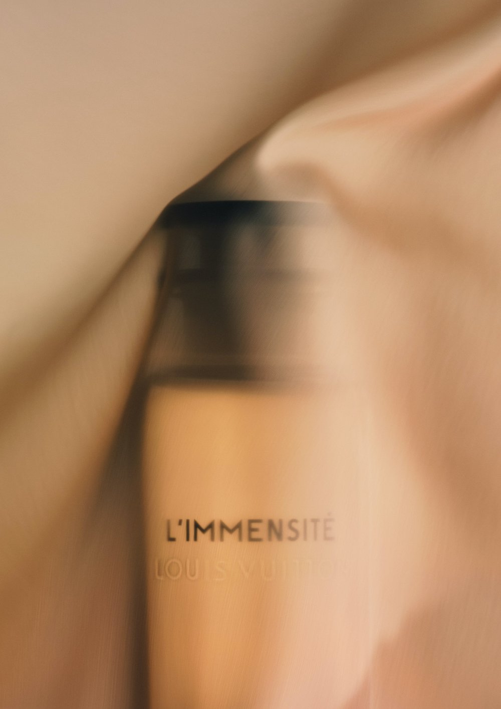 Beige perfume bottle with a dark cap, partially covered in soft beige fabric, creating a blurred and elegant cosmetic presentation.