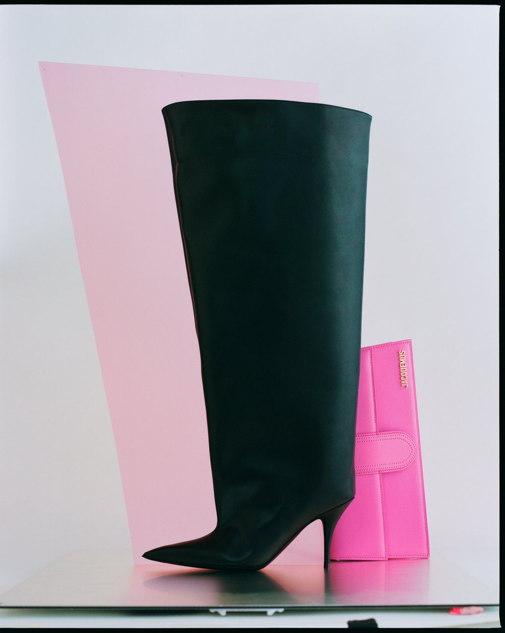 High-heeled black leather boot and pink clutch on a reflective surface with a pink geometric shape against a white backdrop.