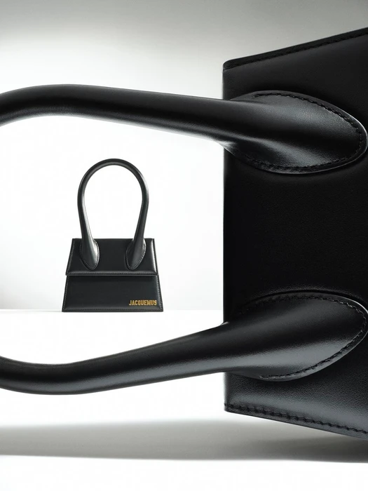 Close-up of a black leather handbag handle framing a smaller black handbag in the background against a white backdrop.