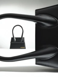 Close-up of a black leather handbag handle framing a smaller black handbag in the background against a white backdrop.