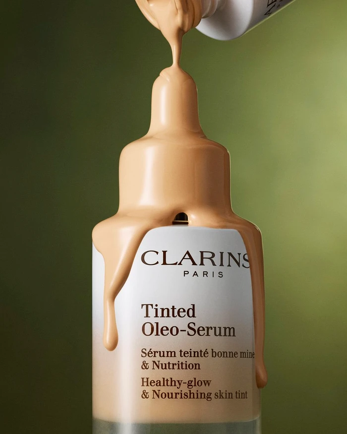 Liquid foundation product shot with tinted serum pouring onto a Clarins glass bottle against a green background