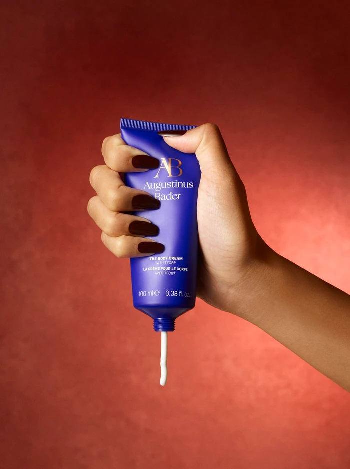 A hand squeezing a blue tube of Augustinus Bader body cream dispensing white lotion against a warm red textured background.