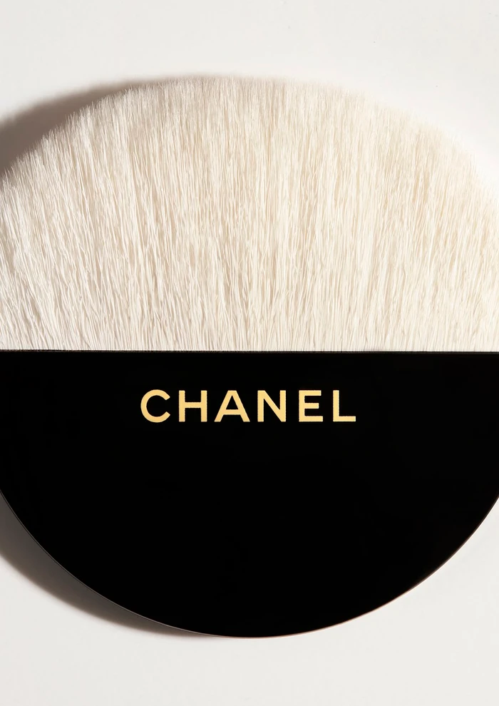 Close-up macro shot of a round makeup brush with soft white bristles and a black handle featuring gold Chanel branding