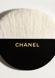 Close-up macro shot of a round makeup brush with soft white bristles and a black handle featuring gold Chanel branding