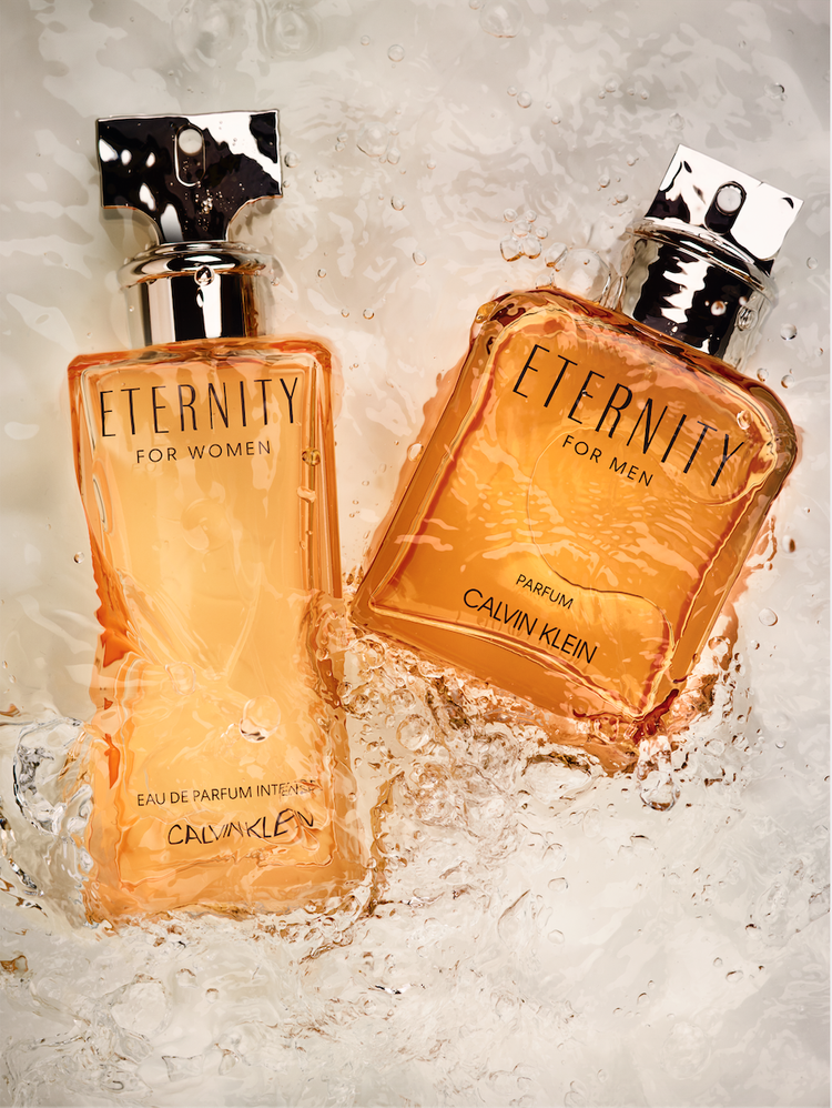 Two Calvin Klein Eternity fragrance bottles, one for women and one for men, are shown partially submerged in water for a beauty shot.