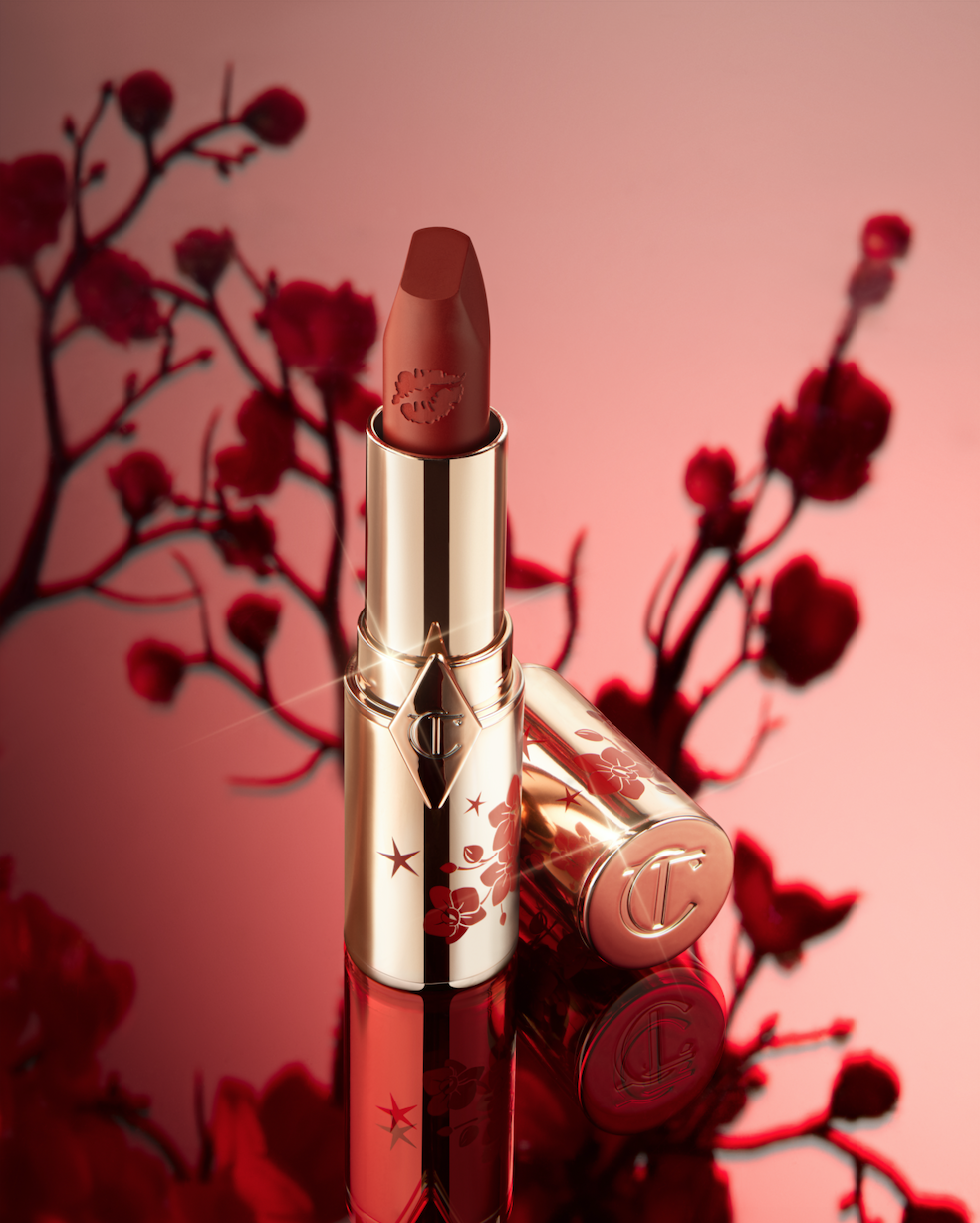 Gold Charlotte Tilbury lipstick sits against a soft pink background with floral accents, showcasing beauty and elegance in product photography.
