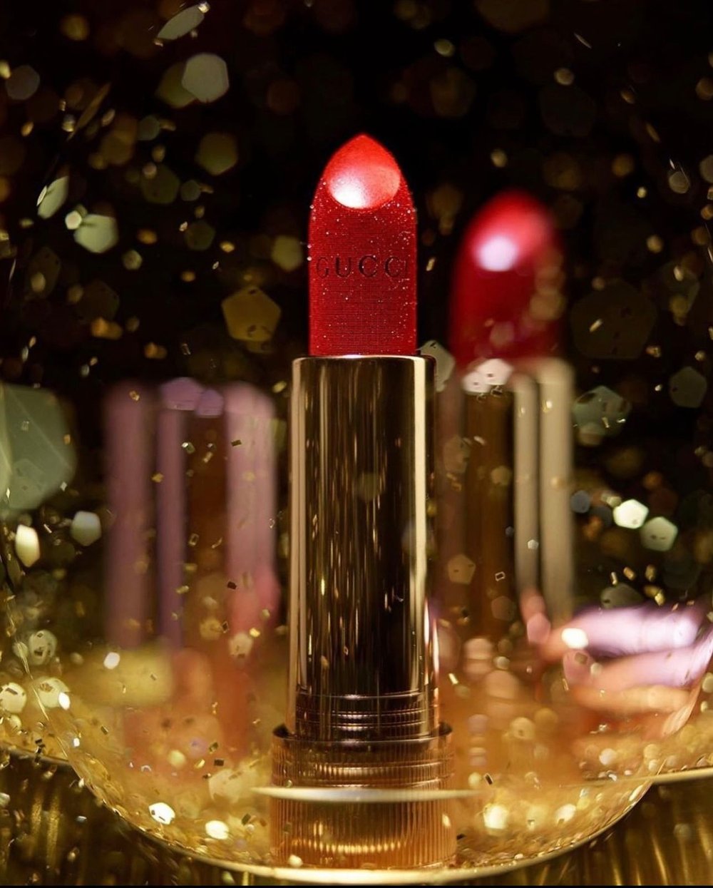 Close-up studio shot of red Gucci lipstick with gold casing surrounded by blurred gold confetti, beauty and fashion photography.