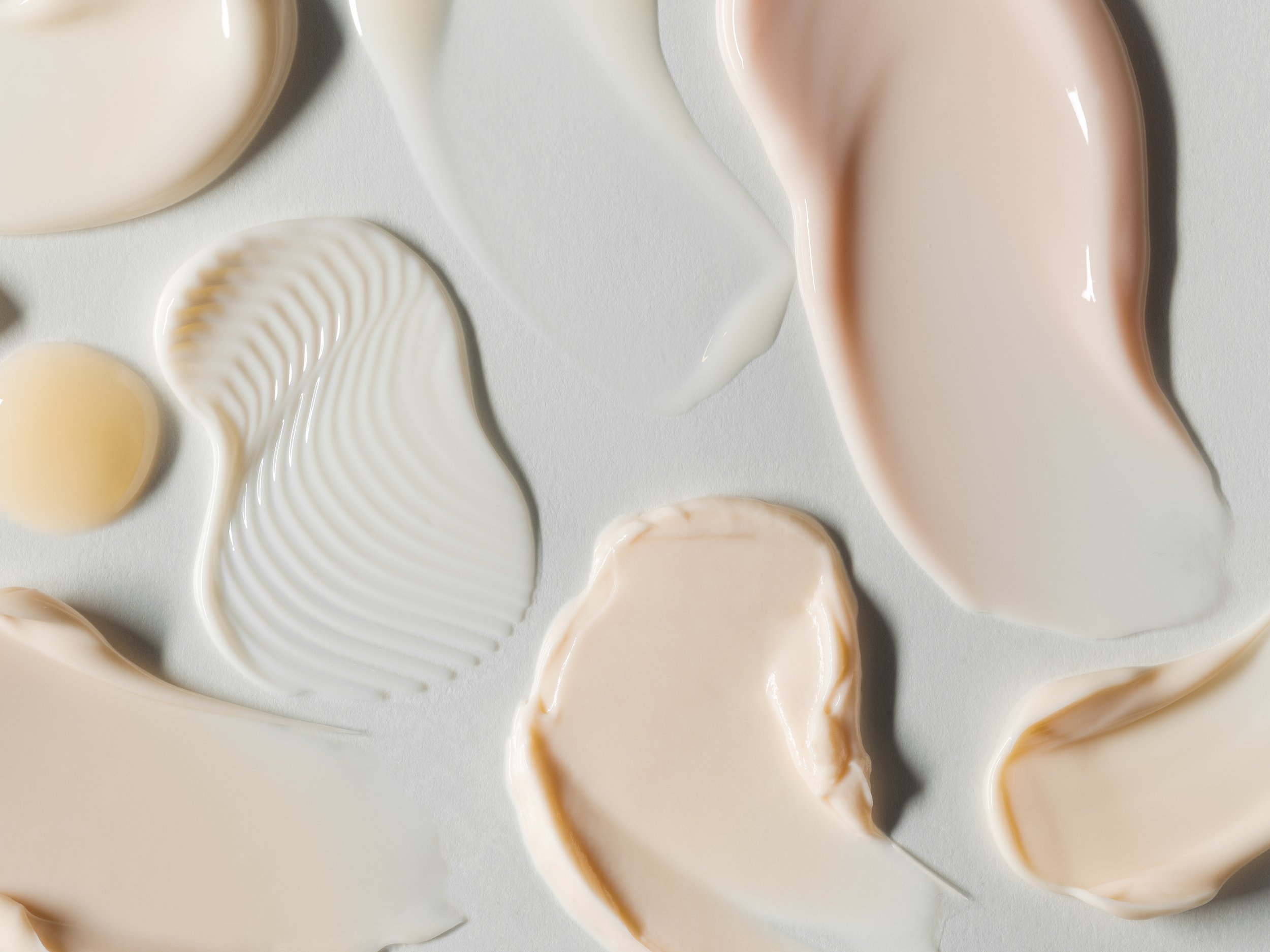 Overhead shot displaying assorted cosmetic cream and lotion smears in shades of white, beige, and peach against a white background.