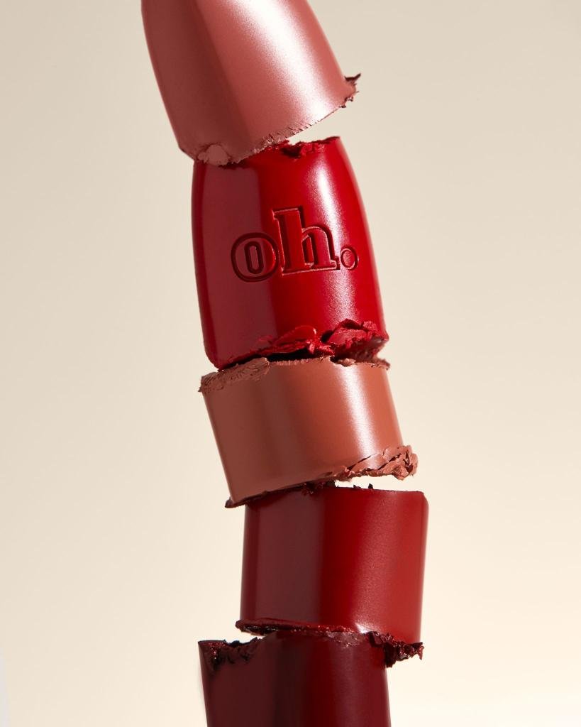 Tower of broken lipstick pieces in varying shades of red and brown against a light beige backdrop, showcasing beauty product.