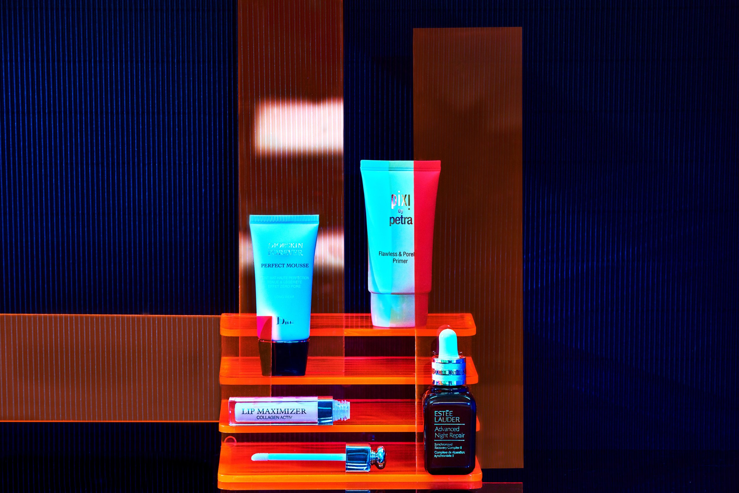 Close-up of Dior, Petra, and Estee Lauder skincare products on an orange shelf against a background with blue and brown stripes.