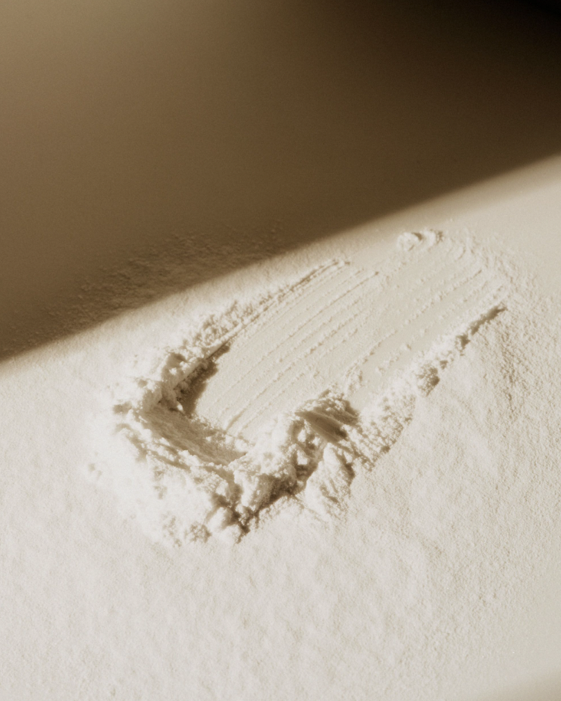High-angle shot of a pile of white powder with linear patterns under stark lighting against a neutral background, creating a minimalist composition.