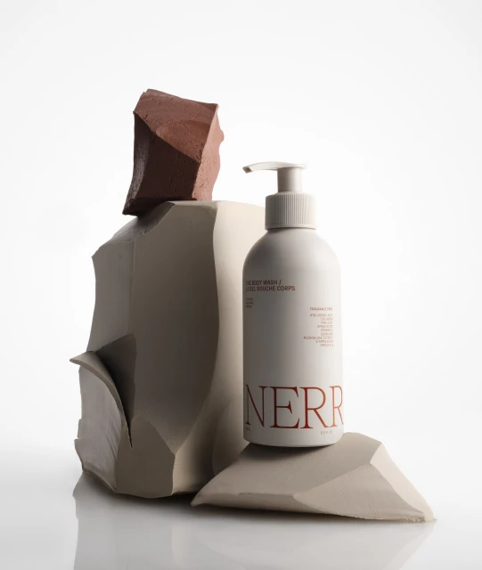 Bottle of body wash with pump dispenser next to beige and brown sculptural stones on a white reflective surface in a studio.
