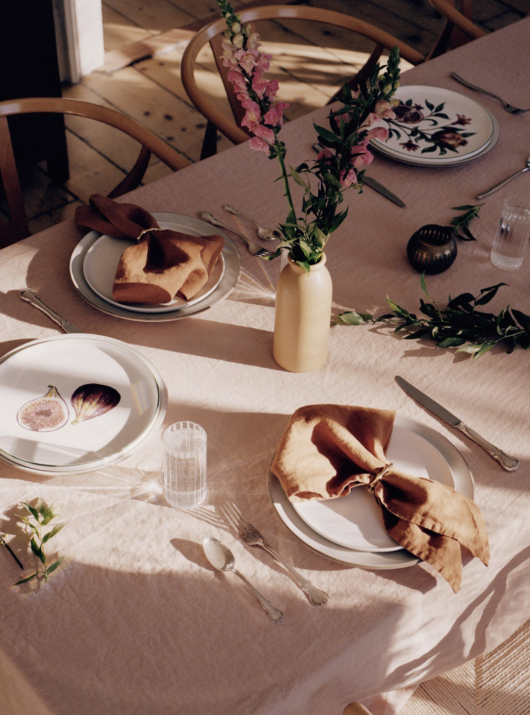 Overhead shot of a table setting with plates, napkins, silverware, flowers, and glasses on a pink linen tablecloth indoors.