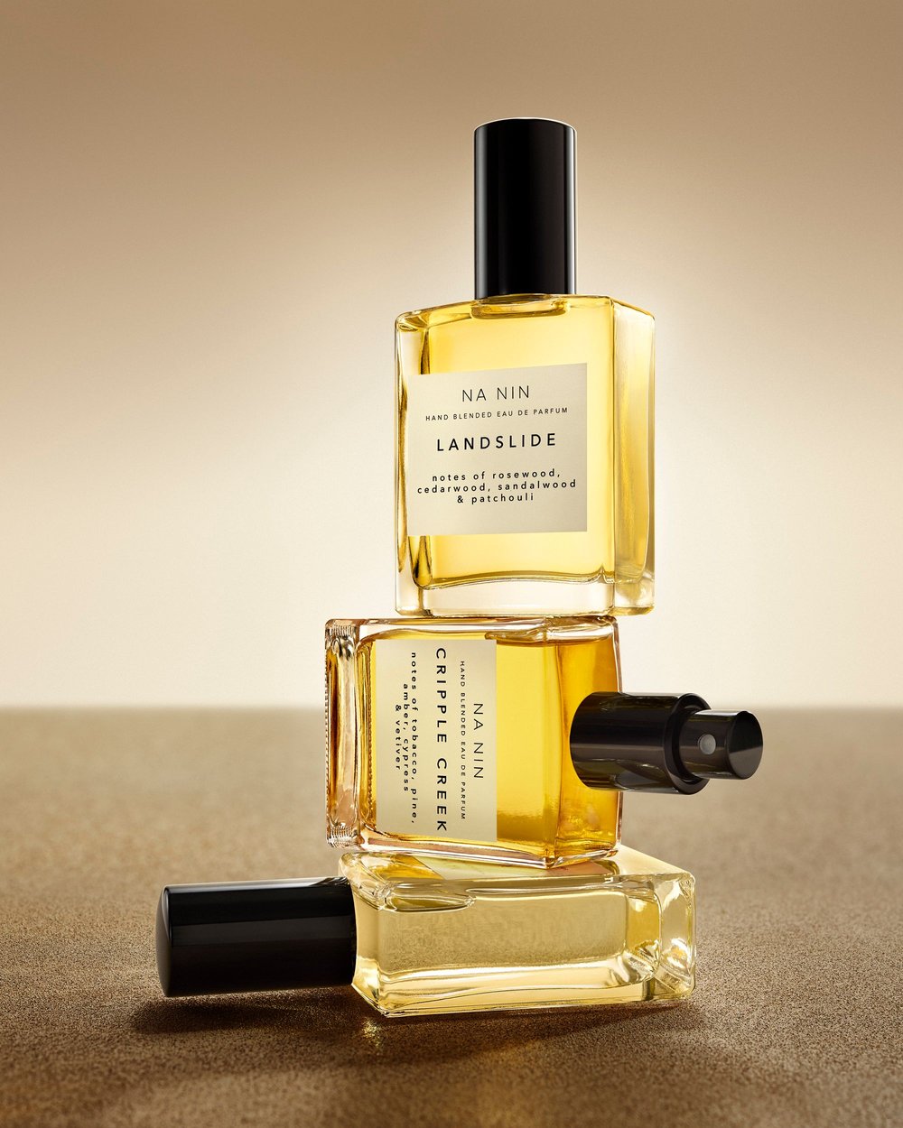 Stacked clear glass perfume bottles with black caps against a gradient beige background, product photography for fragrance brand.