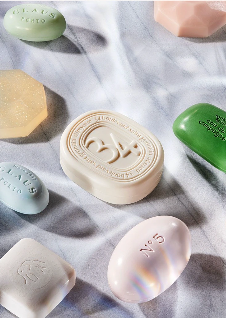 An overhead still life of colorful scented soap bars arranged on a white marble surface with soft natural lighting and shadows.