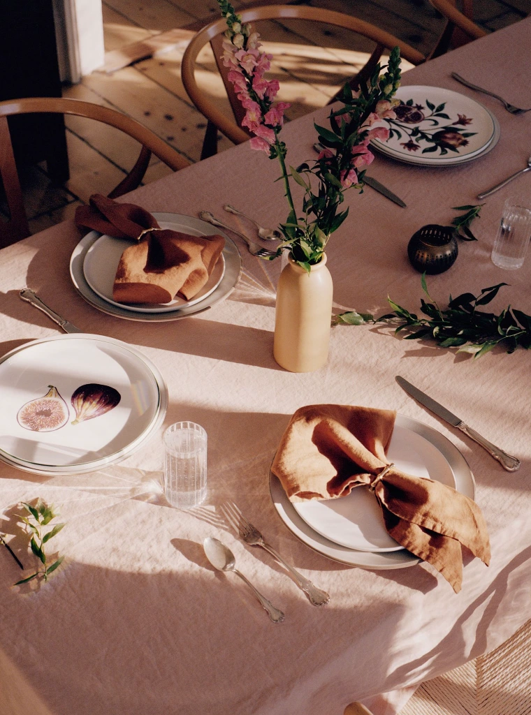 Set-designed dining table with pink linen tablecloth, ceramic vase of pink snapdragons, white fig plates, and rust napkins.